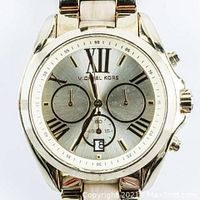 Front face view of the goldtone Michael Kors men's wristwatch showing Roman numerals, three subdials, date window, and brand name.