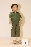 Full front view of the porcelain doll dressed in green sweater and pants, standing on a doll stand.