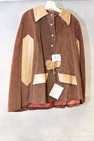Front view of brown suede cape with light brown leather collar, pockets, and sleeve patches. Button front with decorative leather bow detail.