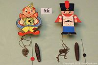 Front view of both clown and soldier whimsical cuckoo clocks, showing full design, weights, and pendulum rods.