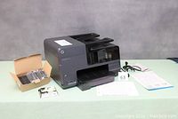 HP Officejet Pro 8610 printer with toner cartridges box, power cord, and documents on table.