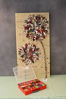 Full view of the decorative button art piece with two large fabric and button flowers mounted on wooden panel, accompanied by red storage bin with 18 compartments of sorted buttons.