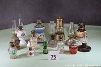 Full lot view showing 14 miniature kerosene oil lamps in various styles, materials, and colors including porcelain, glass, metal, and pewter. Varied chimneys apparent.