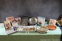 Photo of the entire cooking and baking lot items arranged on a table including multiple cookbooks, various pans and bakeware, measuring cups, and trays.
