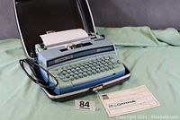 Full view of the blue Smith Corona Coronet Super 10 electric typewriter inside its carrying case with power cord, white paper loaded, and Coronamatic instruction sheet nearby.