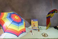 Colorful rainbow umbrella, small bird figurines, and box of inflatable parrot displayed on table.