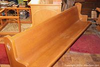 Full view of long oak church pew with carved grape end and plain other end in a room with wood and rugs.