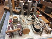 Wide shot of the folk art piece showing the full base with two cows yoked, wood logs, multiple standing tree trunks, a fire pit with cooking pot, and two wooden figures.