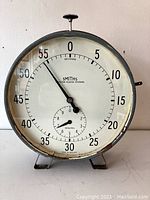 Front view of the large circular metal timer showing the full white dial with seconds and minutes markers and black hands. The Smiths English Clock Systems logo is visible in the center. The timer stands on two metal feet with a knob on top.