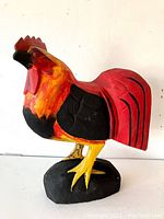 Front and side views of a large hand-carved wooden rooster painted in bright red, orange, yellow, and black colors, standing on a black base.