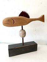 Full view of the folk art fish sculpture showing the carved wooden fish, metal rod, stone, and wooden base.