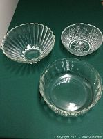 Three large glass bowls on green surface. Top left bowl has vertical ribbed texture. Top right bowl is cut glass with starburst pattern inside. Bottom bowl is clear glass with fluted edges.