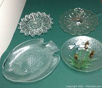 Four clear glass platters and plates arranged on a green surface: fish-shaped platter, cattail plate with handle, star-shaped plate, and another handled plate