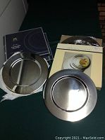 Four stainless steel charger plates with brushed finish, partially stacked, with packaging boxes showing image and brand.