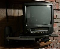 Sansui 9-inch CRT TV with VHS player showing front view on wall-mounted gray stand with shadowed brick and wooden wall background.