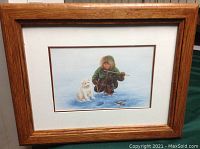 Framed lithograph showing child in winter gear ice fishing with a small white dog, wood frame with white mat and brown line accent