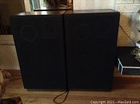 Pair of wood-sided speakers with black fabric front covers and cables on wooden floor.