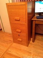 Closed view of oak filing cabinet with three drawers and wooden handles on wood floor.