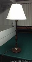 Photo of a 31-inch tall Mid Century Colonial table lamp with a wooden body, brass accents, and a white lampshade placed on a green surface.