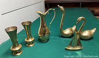 Photo showing two brass swan figurines, a brass sailboat figurine, a brass jug, and two brass vases on a dark surface with white wall background.
