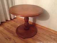 Photo of a round maple wood end/side table with a single turned pedestal base and round bottom support, placed on a wooden floor next to a curtain and wall.