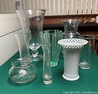 Seven assorted vases of varying shapes and sizes on a green surface near vertical blinds. Includes clear glass vases of different designs and one white ceramic lattice rim vase.
