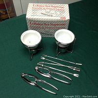 Photo of complete lobster set including two candle-heated butter pots with white ceramic bowls and stands, four metal lobster forks, and two metal lobster shell crackers laid out with box included.