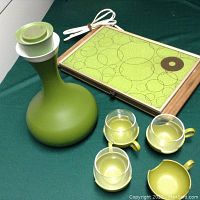 Photo shows a vintage olive green genie bottle with white stopper, a rectangular food warmer with avocado green circular patterned tile top, three glass cups with matching avocado green handle holders, and one empty avocado green cup holder on a dark green surface.