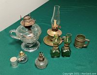 Photo displays six assorted vintage oil lamps arranged on green felt surface featuring different materials and sizes.