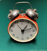 Front view of vintage Orel twin bell alarm clock showing round white face with black numbers 1-12, black hour and minute hands, red second hand, orange body, and chrome bells with handle on top.