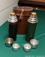 Two vintage metal thermoses with cork tops, two metal cups with and without handles, and a leather carry case on green surface.