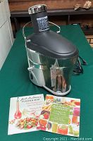 Jack LaLanne Power Juicer with recipe booklets placed on a green surface, showing the main unit and colorful recipe booklet covers.