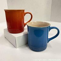 Blue and orange Le Creuset mugs side by side, showing shape and colors, with box beneath.