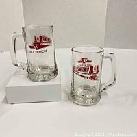 Set of two glass beer steins featuring maroon TTC SRT vehicle graphics and logos, handles visible, one raised on white box for display.