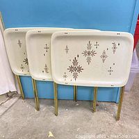 Three vintage folding TV trays standing upright showing metal legs and top trays with brown atomic or firework pattern on white background.