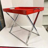 Red lacquer side tray table with chrome X-shaped folding legs shown standing with the tray in place.