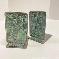 Two metal decorative bookends side by side showing embossed figures and patterns with greenish-blue patina on rectangular vertical panels.