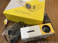 LED projector with yellow and white casing, power adapter, AV cables, and remote control visible, with the box in the background.