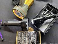 Photo showing large yellow handheld spotlight, black multi-LED round flashlight, and smaller black tactical flashlight in foam-lined box