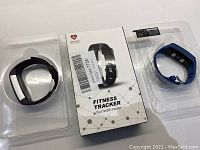 Three fitness tracker watches including one in packaging, two outside packaging on a white surface, one with black band, one blue band.