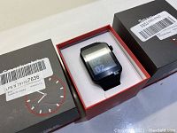 Boxed black smart watch with digital display showing the time 02:02, silicone strap visible, product packaging labeled with barcodes and clock graphics.