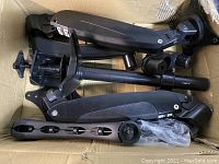 Box containing disassembled black adjustable monitor mount components including arms, clamps, and mounting poles