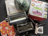 Wide view showing pasta maker with accessories, Starfrit food processor box, digital kitchen scale box, and floral oven mitt.