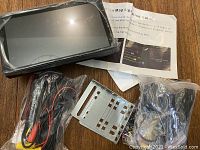 Photo showing the Android touchscreen navigation device, all accessories in plastic bags, various cables, and the metal mounting bracket laid out on a wooden surface.