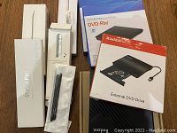 Eight items including stylus pens in boxes and external DVD drives with packaging placed on wooden surface.