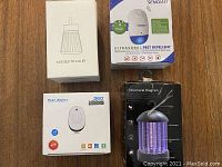 Four pest control items displayed on a wooden surface: a boxed mosquito killer, boxed Pest Reject 360 ultrasonic repeller, Senqiao ultrasonic pest repeller box, and a black boxed ultraviolet mosquito killer lamp.