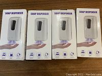 Four boxed white soap dispensers lined up on a table with front of boxes showing product images and icons for waterproof and contactless operation.