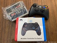 Lot with two vintage NES style wired controllers, black Xbox 360 wireless controller, and boxed Nintendo Switch wireless controller on wood surface.