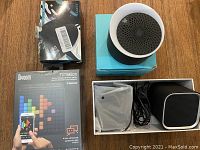 Photo showing 4 Bluetooth speakers and their boxes laid on a wooden surface: Divoom TimeBox box, a compact cylindrical speaker on a blue box, and two small square speakers in white boxes with cables.