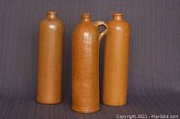 Three ceramic wine/spirit bottles standing upright showing height and general form. Bottles are brown-glazed and cylindrically shaped, one with handle.